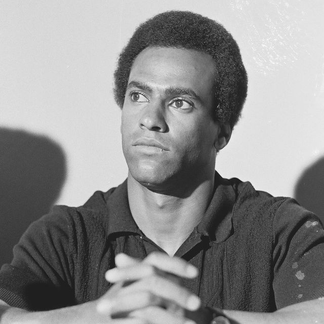 huey p newton sits with his hands clasped in front of him, he looks to the left and wears a dark collared shirt