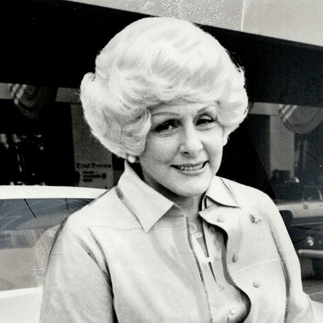 mary kay ash smiles at the camera, she wears a collared coat and large button earrings