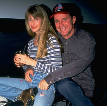 brynn omdahl and phil hartman smile at the camera while seated, both wear jeans and long sleeve shirts, hartman also wears a baseball cap and embraces brynn