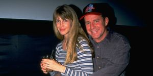 brynn omdahl and phil hartman smile at the camera while seated, both wear jeans and long sleeve shirts, hartman also wears a baseball cap and embraces brynn