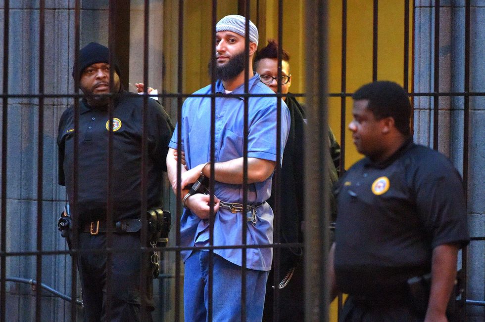 adnan syed wears a blue prison outfit, a gray cap, and handcuffs, he is looking toward the camera from behind bars and three officers are in the vicinity