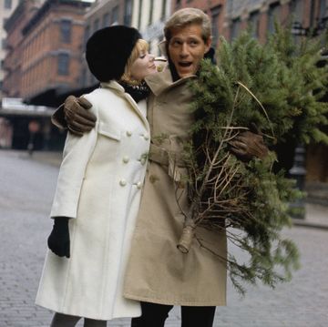 george segal and maggie london walk down a city street, their arms wrapped around each other, he carries a small christmas tree she wears a white wool coat by dani jrs, black and white muffler by einier accessories, black bubble hat by veaumont, black gloves by hansen, and grey stockings by berkshire he wears a khaki trench coat with slacks photo by louis faurercondé nast via getty images
