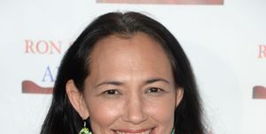 irene bedard smiles at the camera while standing in front of a white backdrop, she wears a black sequin top, a silver chain necklace, and colorful dangling earrings