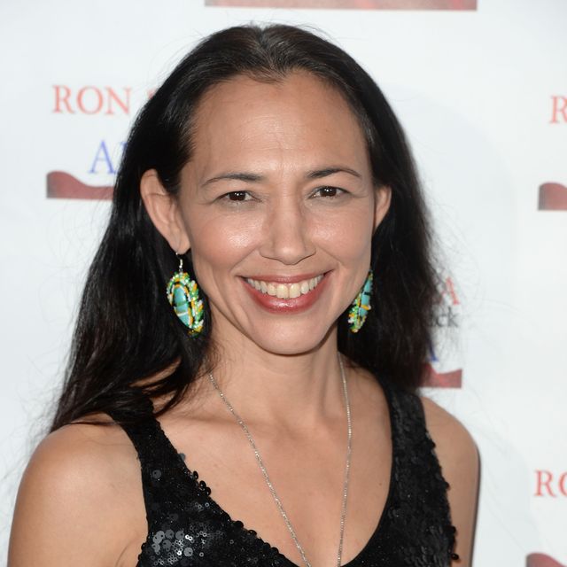 irene bedard smiles at the camera while standing in front of a white backdrop, she wears a black sequin top, a silver chain necklace, and colorful dangling earrings