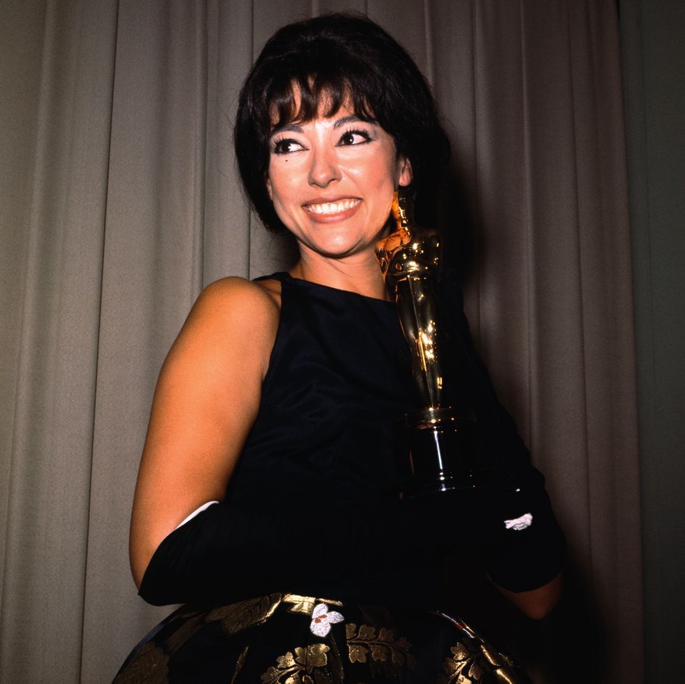 rita moreno smiles and holds an academy award statuette, she wears a black dress and long black gloves