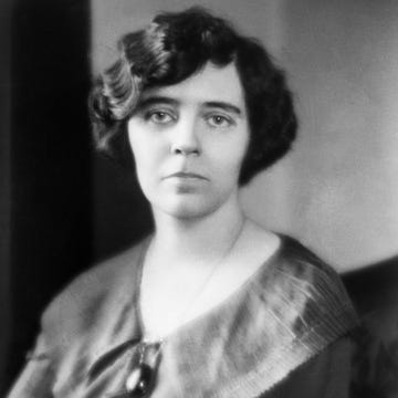 a woman stares at the camera, she wears a large pendant necklace and a blouse with a wide collar