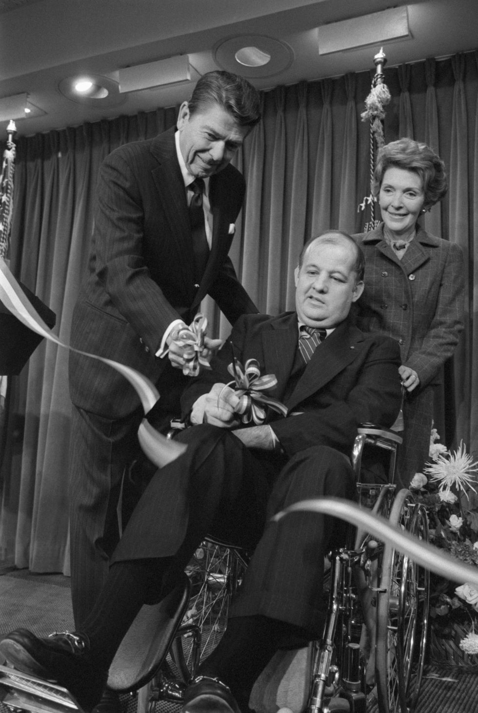Press Secretary Jim Brady, with Ronald and Nancy Reagan, cuts the ribbon opening the renovated press center at the White House in November 1981