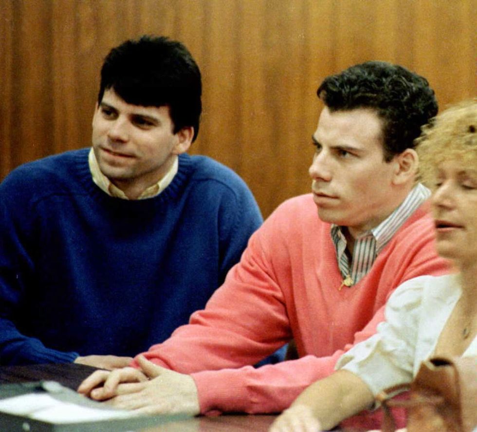 lyle and erik menendez sit at a table inside a courtroom, lyle is wearing a royal blue sweater on top of a yellow collared shirt, and erik is wearing a pink v neck sweater on top of a striped collared shirt