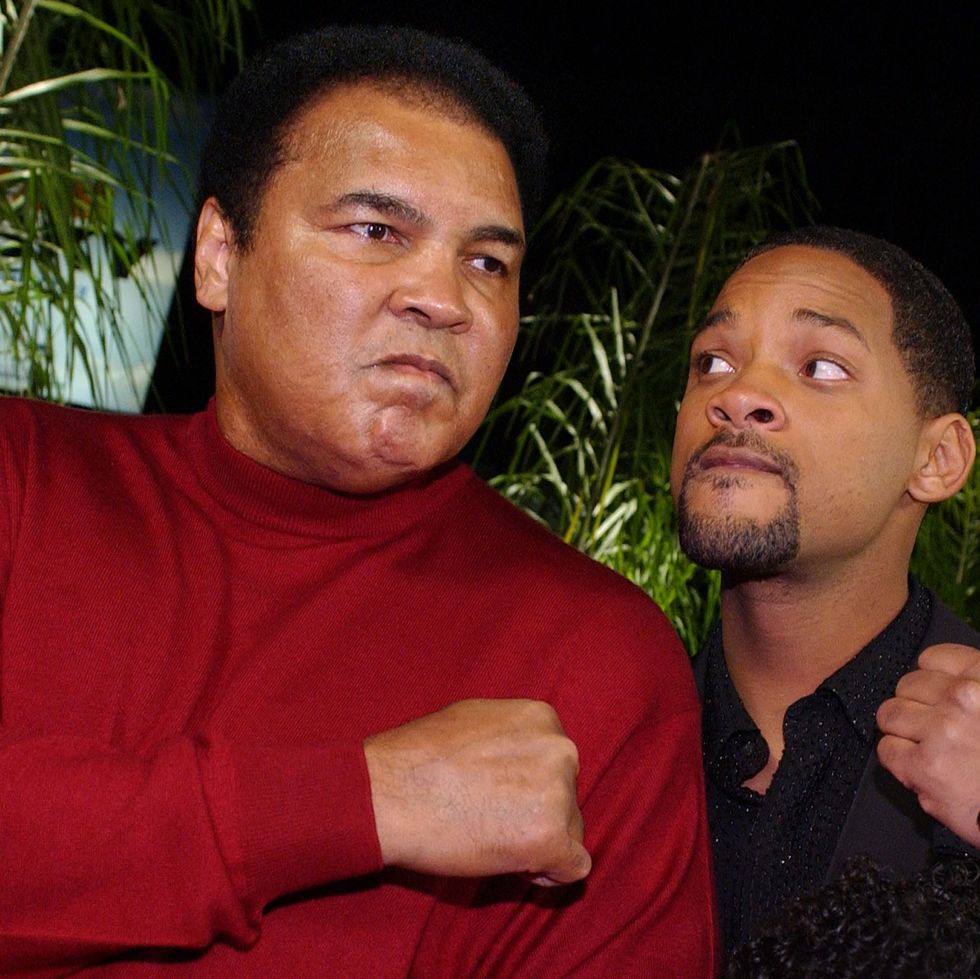 muhammad ali and will smith pose for a photo with each holding one fist up on their chest, ali wears a red long sleeved shirt and smith wears a black shirt