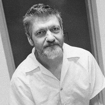 ted kaczynski looks at the camera with a neutral expression, he is wearing a white collared button up shirt and stands in front of a doorway