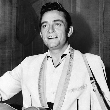 johnny cash smiles as he stands and plays a guitar, he wears a light colored collared shirt and jacket with a patterned lapel