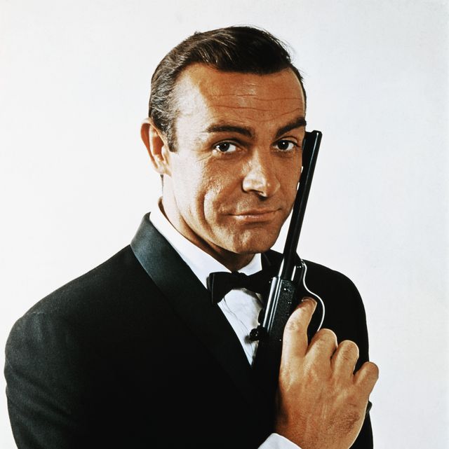 original caption waist up portrait of sean connery, as james bond, caressing the barrel of a gun against the side of his face connery is wearing a tuxedo and bow tie and smiling slightly