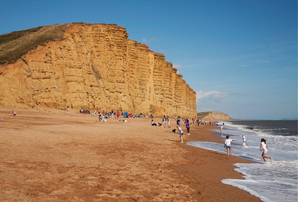 sandstone cliffs and beach, west bay, bridport, dorset, england. the orange stone cliffs stand in front of blue skies and people are dotted across the beach.