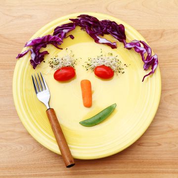 a playful vegetable face adorns a large yellow plate on a wood surface table top the face is made with cherry tomatoes for the eyes, alfalfa sprouts for the eye brows, a baby carrot for the nose and red cabbage for the hair a lopsided smiling mouth is made with a sugar snap pea a fork with a wood handle is also on the plate concept of making it fun to eat your vegetables and enjoy a healthy diet