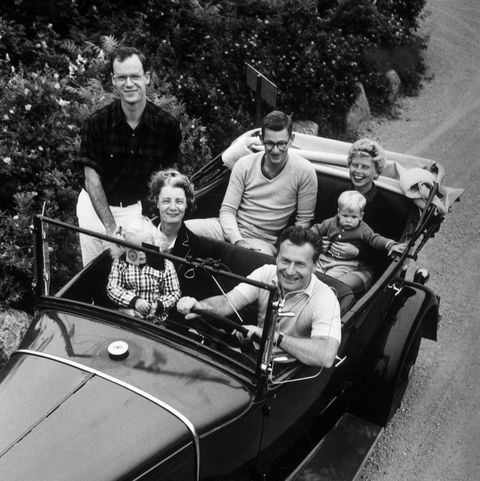 Nelson Rockefeller drives a car with his wife Mary "Tod" Rockefeller seated beside him, holding their granddaughter Meile. Their son Rod is seated in the back with his wife Barbara and son Peter, while his brother Steve stands beside them.