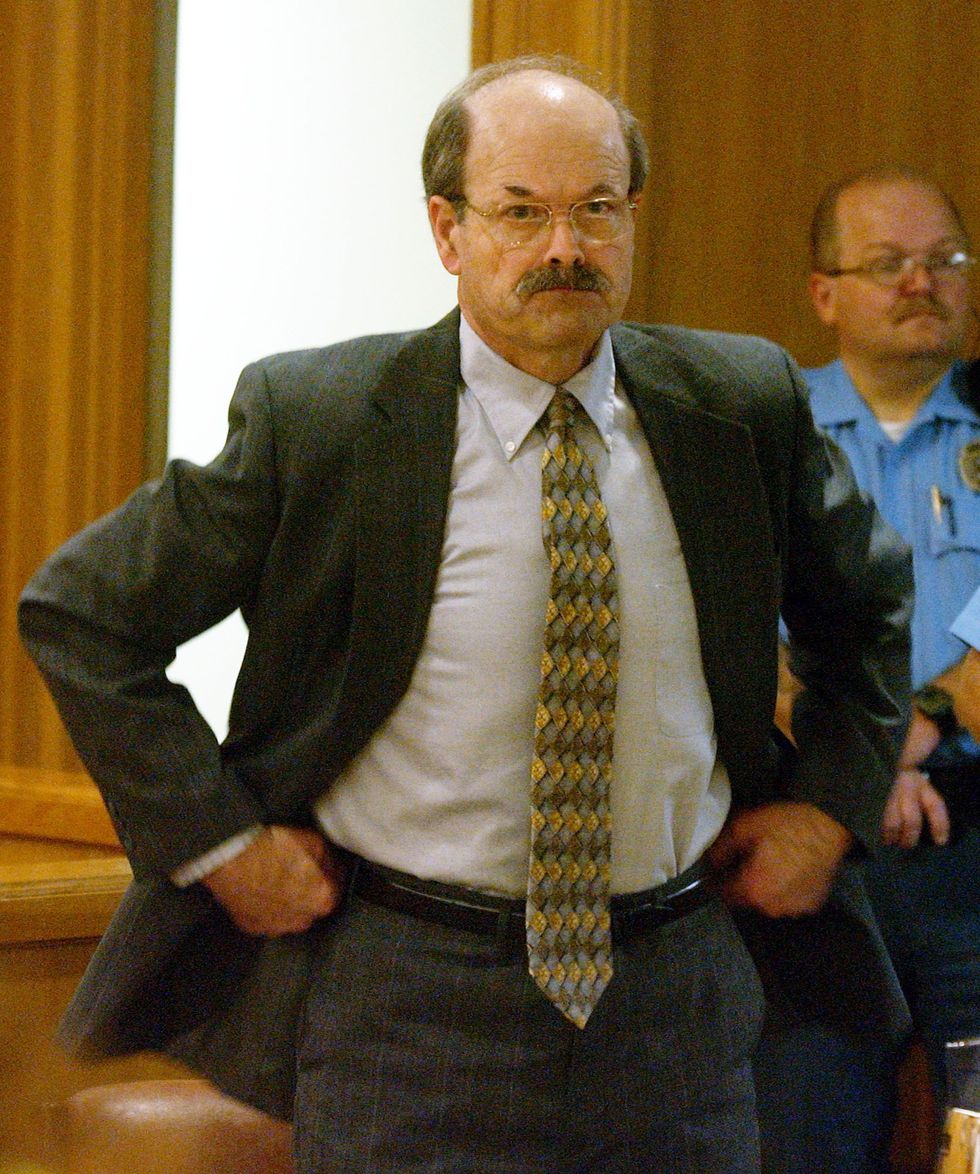 dennis rader pulls at his belt and pants waistband while standing inside a wood paneled room, he wears a gray suit with a white collared shirt and yellow and brown patterned tie, behind him is an officer standing with his hands together