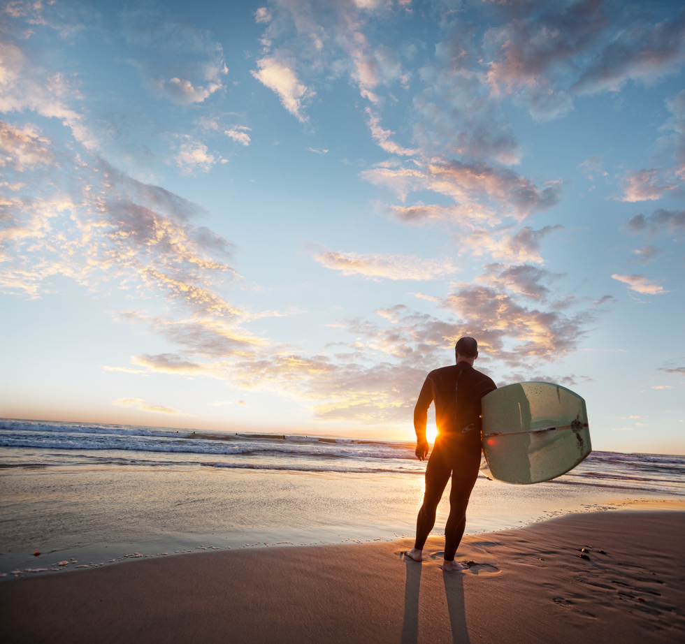middle aged man wearing a wetsuit standing on beach at sunset while holding surfboard at dana point, california, rear camera angle