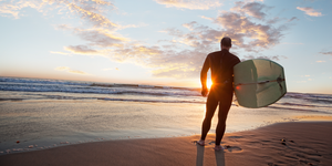 middle aged man wearing a wetsuit standing on beach at sunset while holding surfboard at dana point, california, rear camera angle