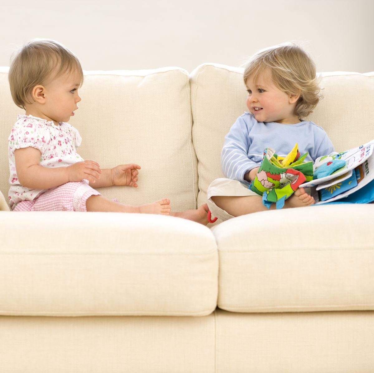 Baby girl (9-12 months) looking at toddler boy (12-15 months) on sofa with toys, side view