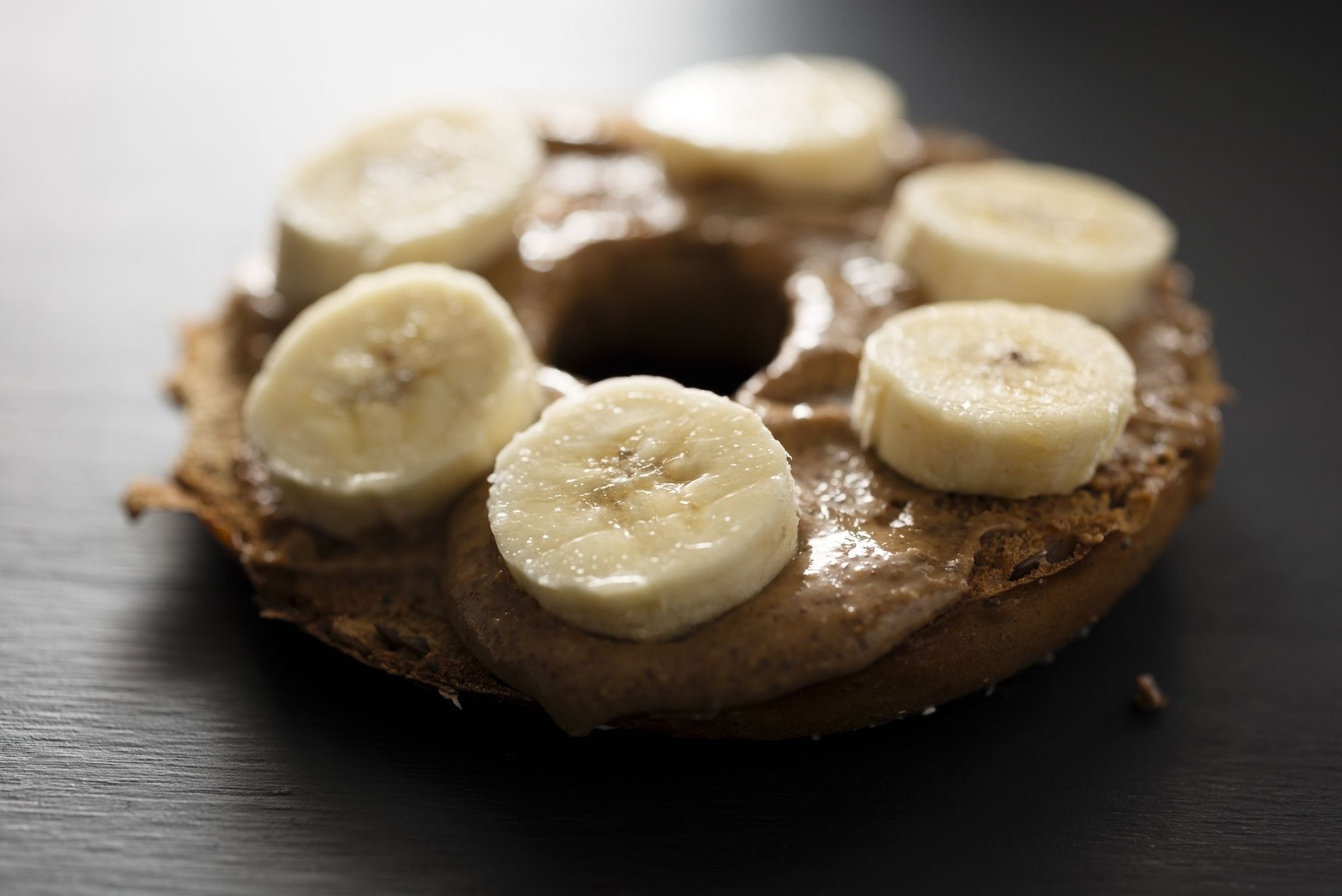 toasted bagel with almond butter spread and sliced banana, backlit with window light on a painted black chalkboard backdrop