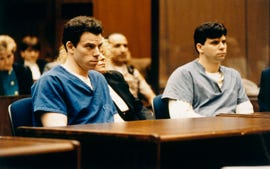 lyle and erik menendez sit inside a courtroom at wooden tables and face forward, lyle is wearing a blue prison uniform, erik is wearing a gray prison uniform with a white long sleeve shirt underneath