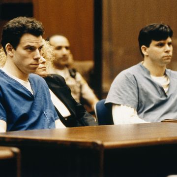 lyle and erik menendez sit inside a courtroom at wooden tables and face forward, lyle is wearing a blue prison uniform, erik is wearing a gray prison uniform with a white long sleeve shirt underneath