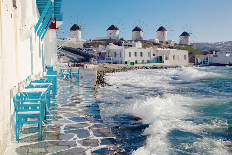 cafe tables and chairs right near the sea with waves overlooking famous mykonos windmills