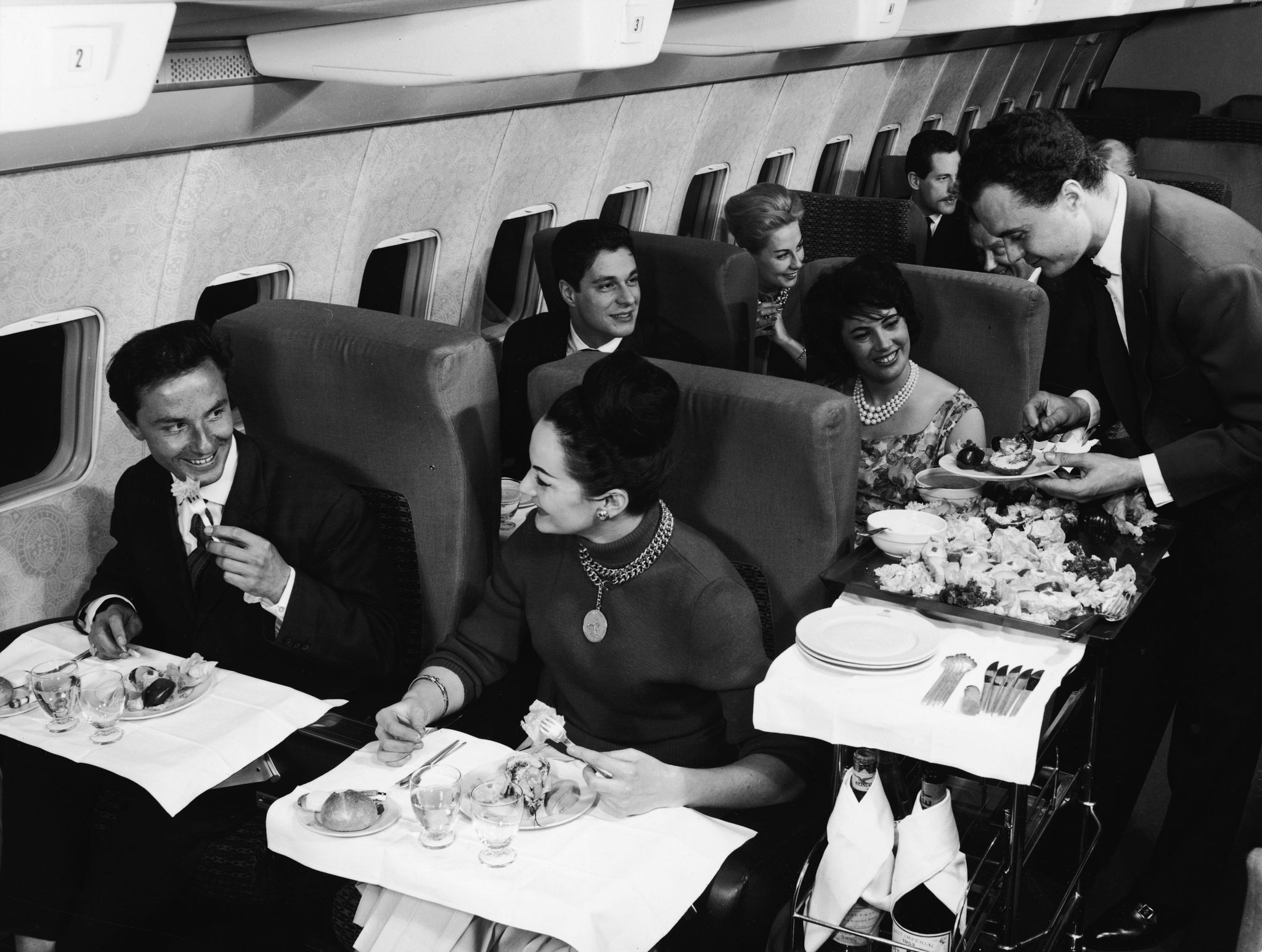 interior view of the first class compartment of a commercial passenger plane a boing shows, in the foreground, a well dressed couple as they smile and enjoy their meal, while behind them a flight attendant in a bow tie serves another happy couple, 1950s photo by authenticated newsgetty images