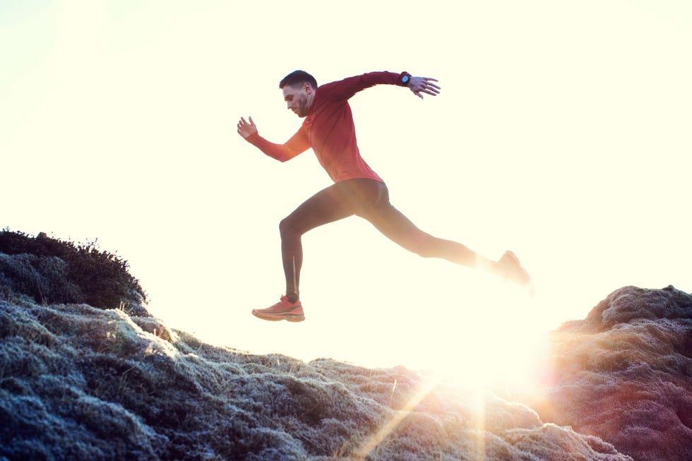 male runner dressed in red top leaps and powers his way though grass hilltop side angle view of runner jumping, golden hue sky