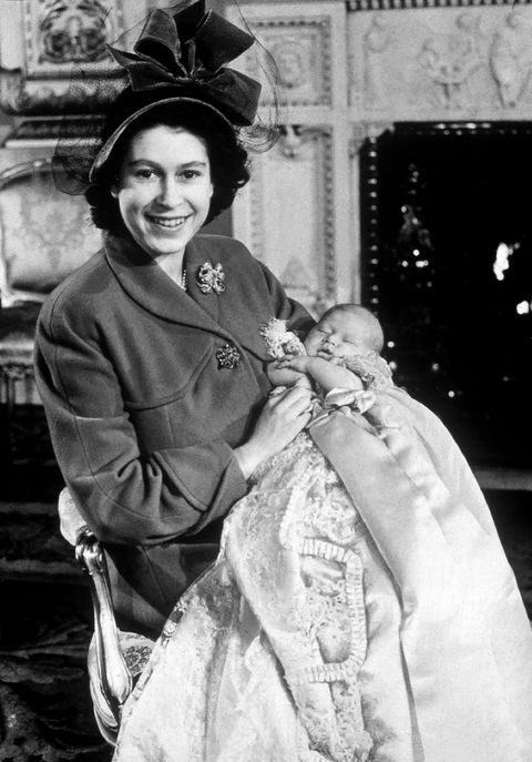 princess elizabeth sits in a chair and holds prince charles as an infant, she is smiling at the camera and wearing an ornate hat and long coat, he is sleeping and wearing a long christening gown