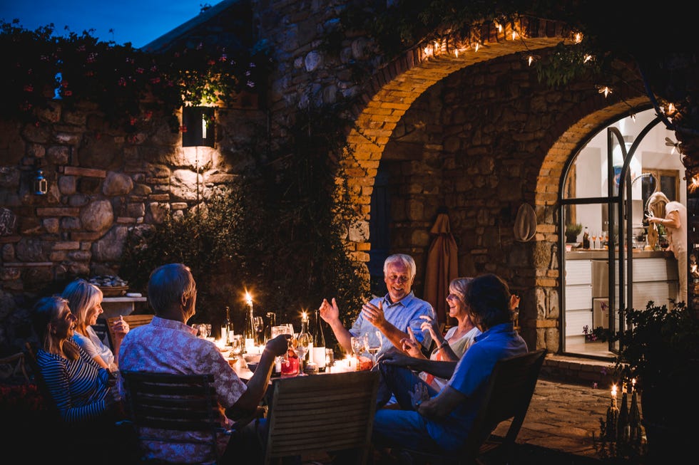 a group of mature friends are sitting around an outdoor dining table, eating and drinking they are all talking happily and enjoying each others company as darkness falls the image has been taken in tuscany, italy