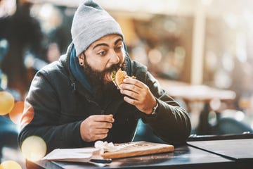 closeup of bearded hipster guy gorging on a burger like if nobodys watching hes sitting outdoors on a cold sunny day hes wearing a dark jacket, gray cap and has long brown beard