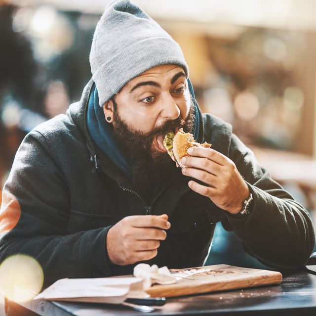 closeup of bearded hipster guy gorging on a burger like if nobodys watching hes sitting outdoors on a cold sunny day hes wearing a dark jacket, gray cap and has long brown beard