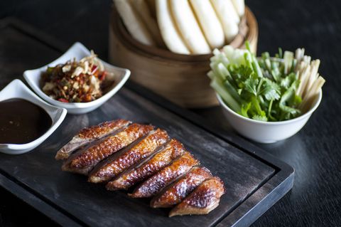roasted sliced chinese style duck breast on a dark wooden chopping board with sweet soy dip and chopped chilli sauce, with asian greens and steamed rice buns on the side