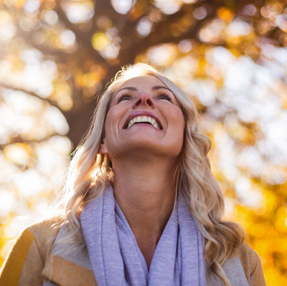smiling woman looking up against trees at park during autumn