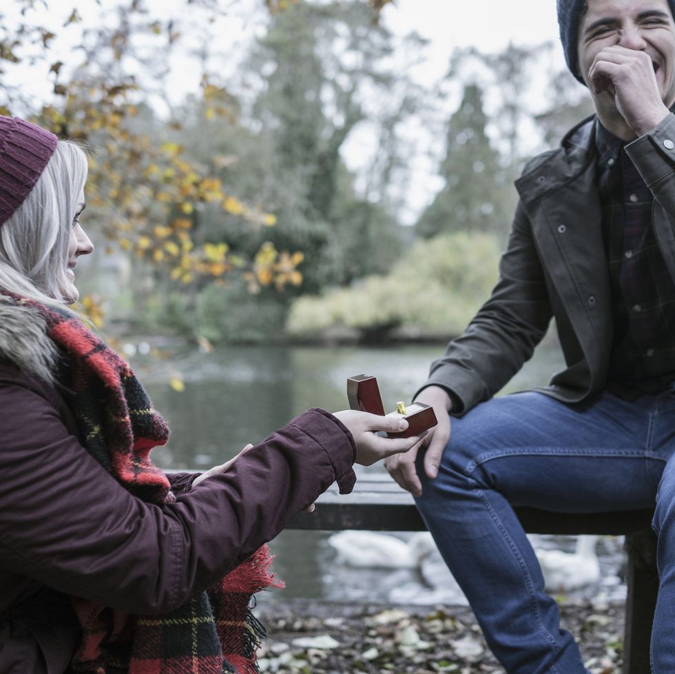 a woman kneeling on one knee proposing to a surprised man on a park bench