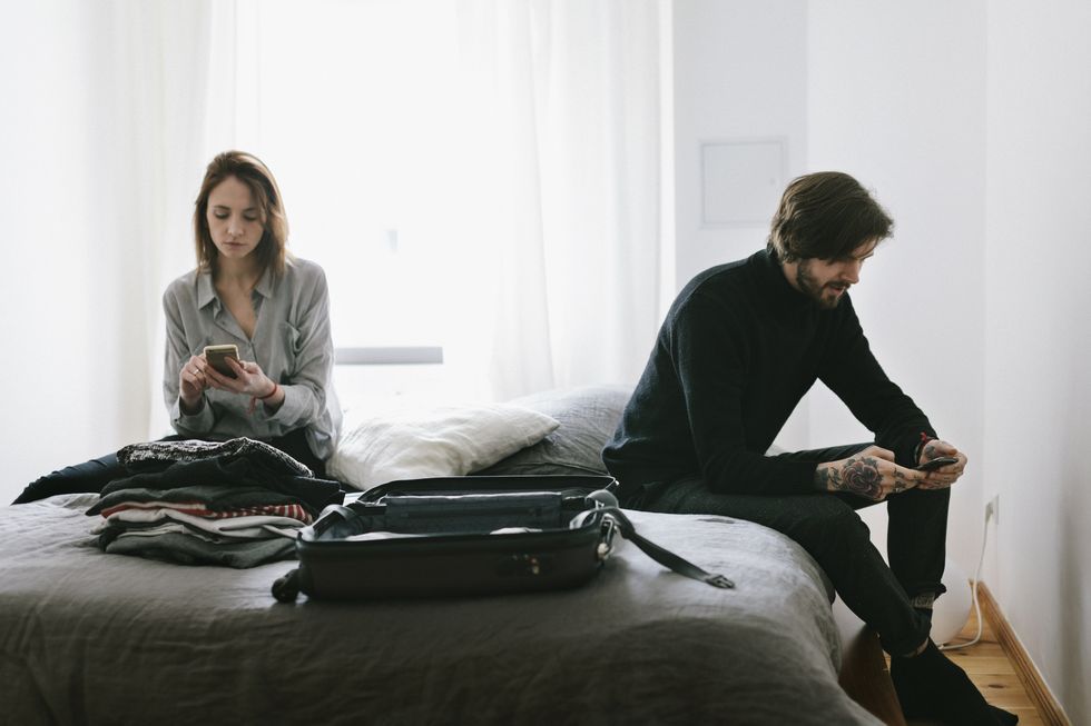 couple sitting on bed checking smartphone messages with unpacked suitcase lying on bed