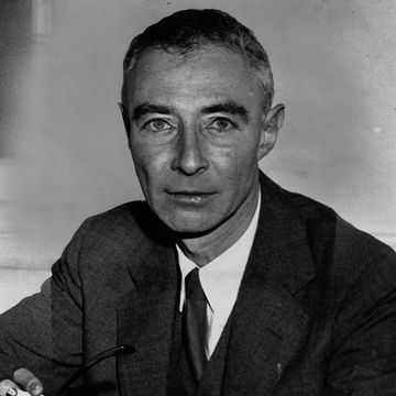 j robert oppenheimer looks at the camera with a neutral expression on his face in a black and white photo, he wears a dark suit with a white collared shirt and dark tie