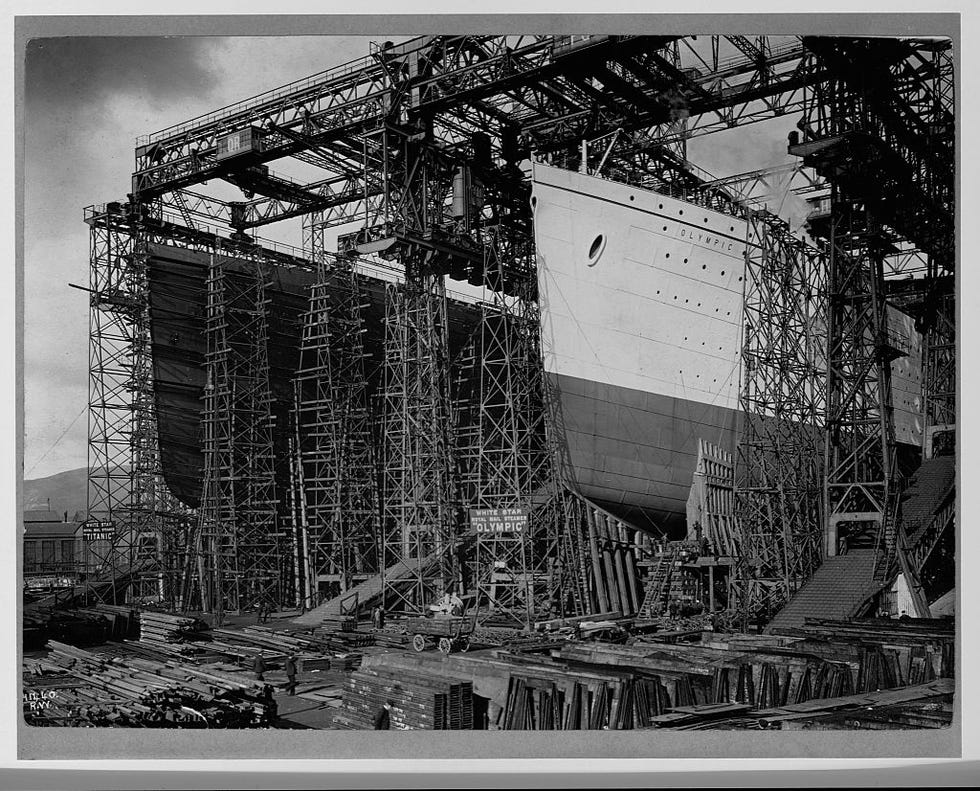 a shipyard where two large ships are being worked on, tall scaffolding surrounds all sides of the ships as construction material appears in the foreground