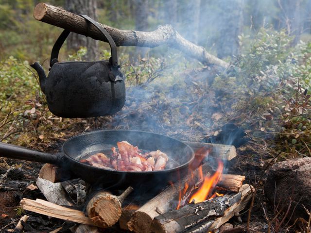 Blackened tin kettle boiling water and pan cooking bacon over flames from campfire.