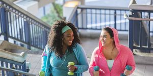 two multi ethnic young women exercising together they are looking at each other, smiling, as they climb a staircase holding hand weights