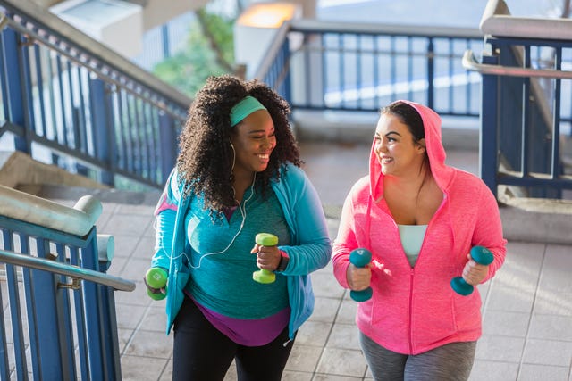 two multi ethnic young women exercising together they are looking at each other, smiling, as they climb a staircase holding hand weights