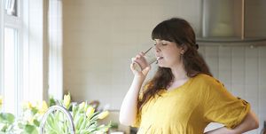 a pregnant woman drinks a glass of water in the kitchen of her home with fresh picked spring flowers in vase