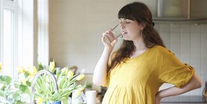 a pregnant woman drinks a glass of water in the kitchen of her home with fresh picked spring flowers in vase