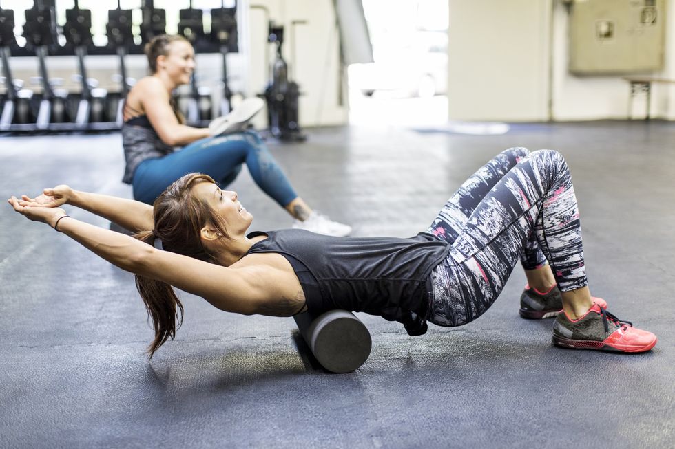 side view of woman rolling on foam roller with friend at health club female athletes are exercising together in gym they are in sports clothing