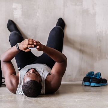 end of training tired african muscular man is lying on floor and looking to his mobile phone he is resting with barefoot legs on wall