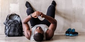 end of training tired african muscular man is lying on floor and looking to his mobile phone he is resting with barefoot legs on wall