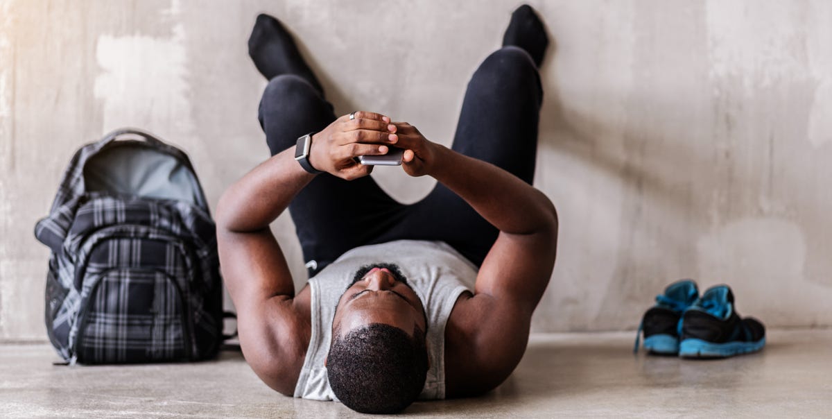 end of training tired african muscular man is lying on floor and looking to his mobile phone he is resting with barefoot legs on wall