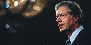 jimmy carter in profile, he is wearing a suit jacket, blue collared shirt and black tie with a blue and white strip, he has a solemn look on his face