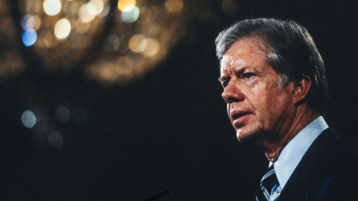 jimmy carter in profile, he is wearing a suit jacket, blue collared shirt and black tie with a blue and white strip, he has a solemn look on his face